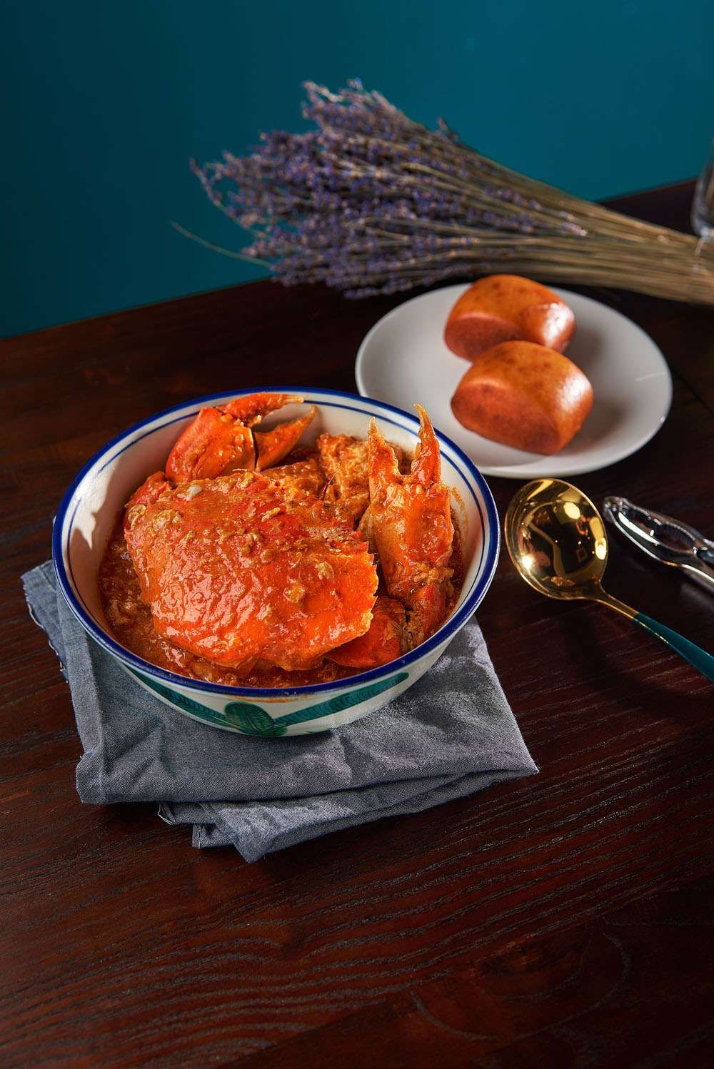 A whole crab coated in red chilli crab sauce in a blue-rimmed bowl with fried mantou beside it.