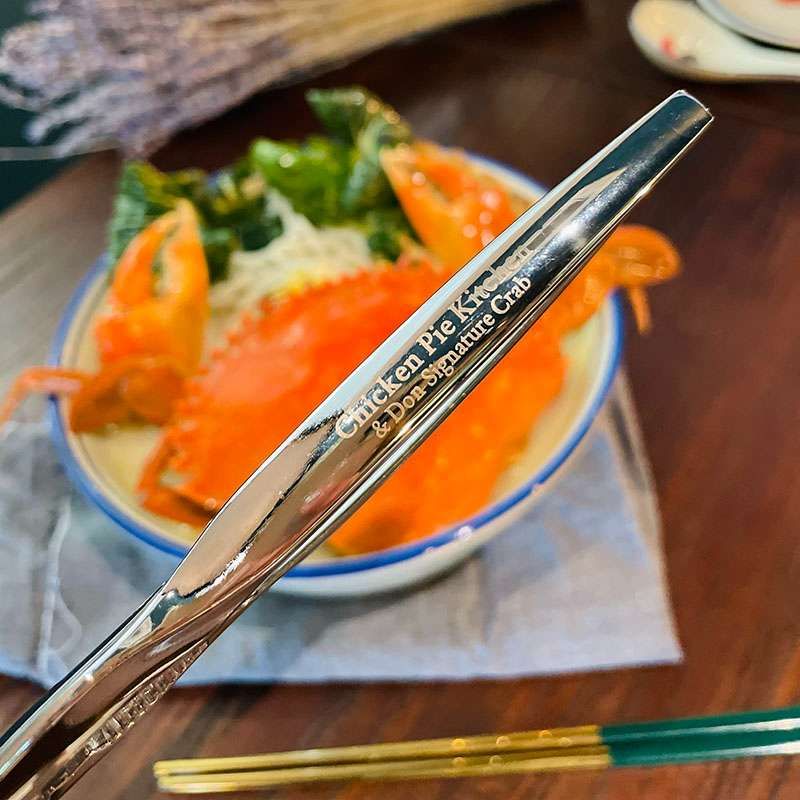 A whole crab in a bowl of creamy laksa with a silver metal crab cracker engraved with the kitchen's name lying across it.