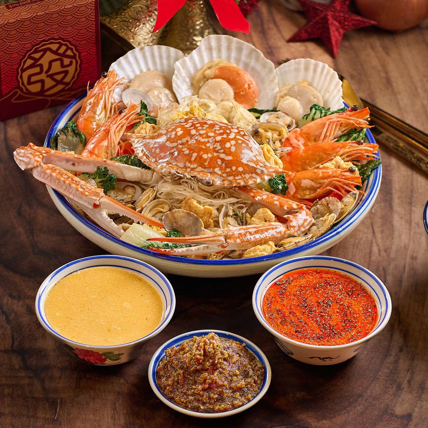 A large bowl of seafood bee hoon topped with a flower crab, scallops in shell and prawns, with three small bowls of sauce in front.