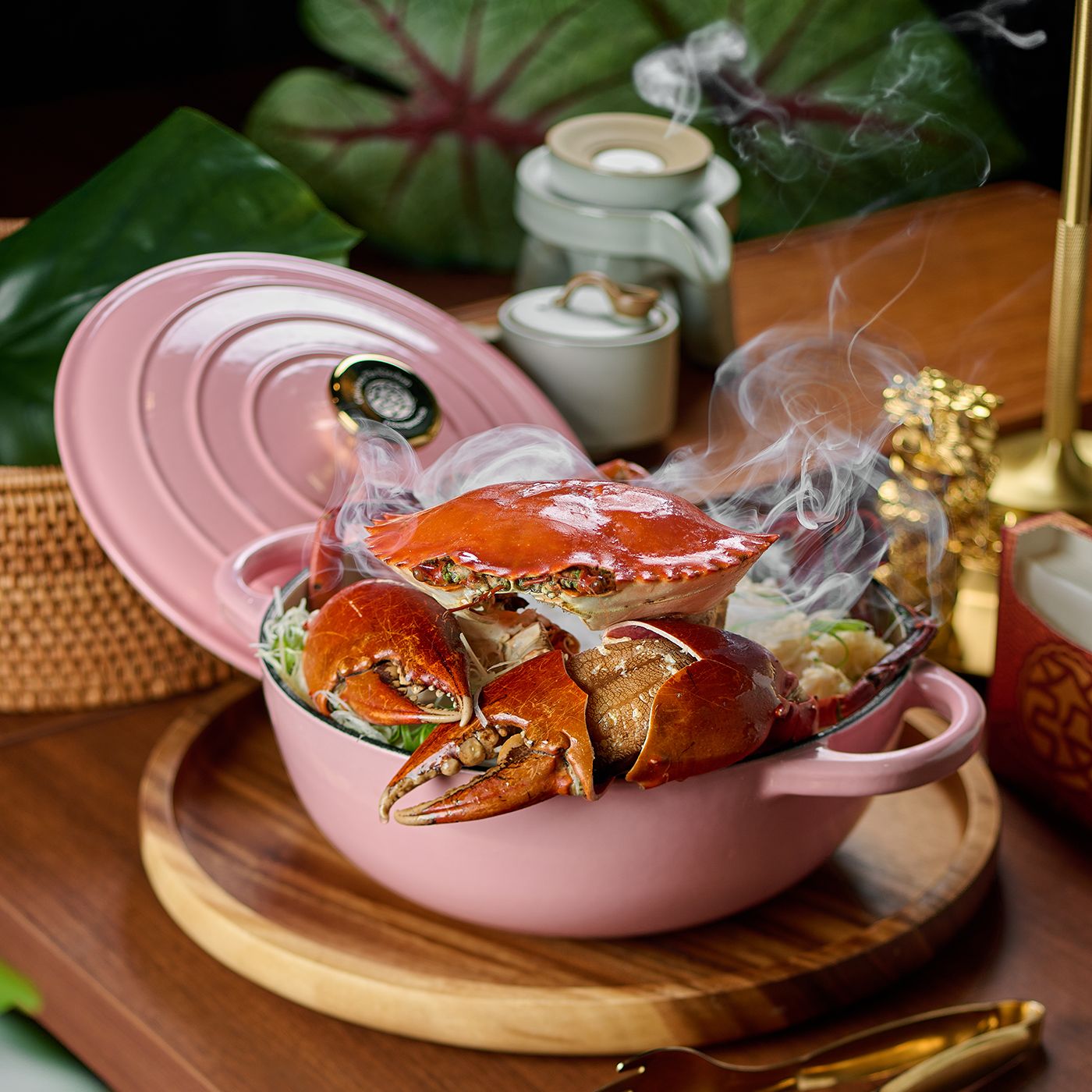 A whole crab with raised claws in a pink cast-iron pot, steaming, on a wooden board beside a glass jar and green leaves.